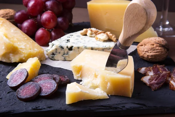 Blandade ostar, nötter, druvor, frukt, rökt kött och ett glas vin på ett serveringsbord. Mörk och lynnig stil. — Stockfoto