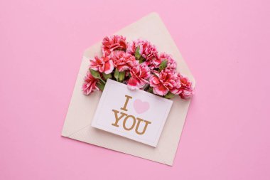 Üzerinde pembe çiçekler ve bir kart olan bir zarf. Pembe arka planda seni seviyorum.