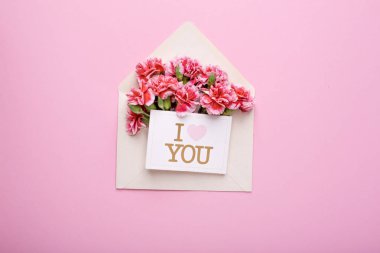 Üzerinde pembe çiçekler ve bir kart olan bir zarf. Pembe arka planda seni seviyorum.
