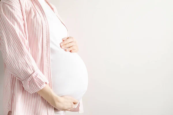 Beyaz tişörtlü hamile bir kadın, elleri telif boşluğu olan hafif bir arka planda karnını tutuyor ve elleriyle karnını tutuyor. Annelik ve sevgi