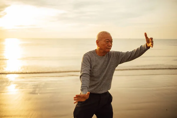 Старший азиатский мужчина практикует тайцзи и йогу позы на пляже — стоковое фото