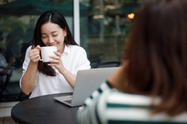 Bilgisayarla çalışan Asyalı kadın kahve dükkanında kahve içiyor, gülümsüyor ve arkadaşlarıyla konuşuyor.