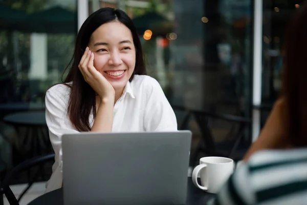 コーヒーショップでパソコンとコーヒーを飲むアジアの女性の笑顔と幸せな顔と友人と話す ストック写真