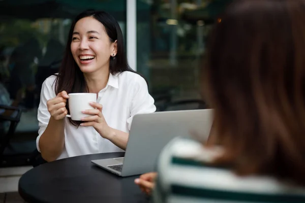 コーヒーショップでパソコンとコーヒーを飲むアジアの女性の笑顔と幸せな顔と友人と話す ストック画像