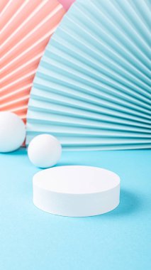 Mavi yüzeye yerleştirilmiş dairesel beyaz kaide renkli kağıt yelpaze ve kürelerle çevrili, neşeli ve modern bir ekran yaratıyor, kopya alanı olan hikayeler.