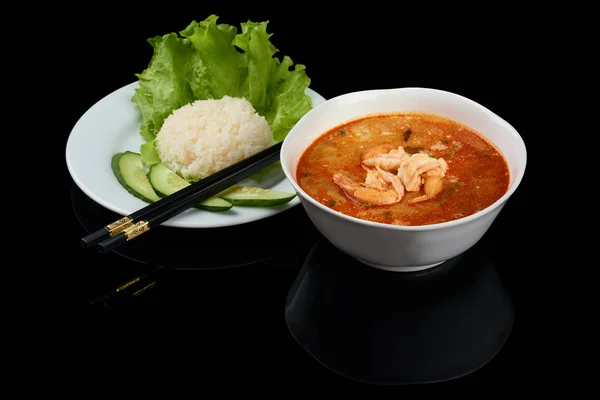 Tom yum or tom yam is a type of hot and sour Thai soup, usually cooked with shrimps. Served with rice and chopsticks. Isolated on black reflective background.