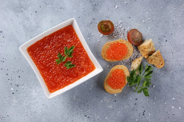 Seafood. Red caviar in a white bowl, sandwiches with caviar, herbs and tomatoes on a light grey background. Flatlay, top view. Background image, copy space