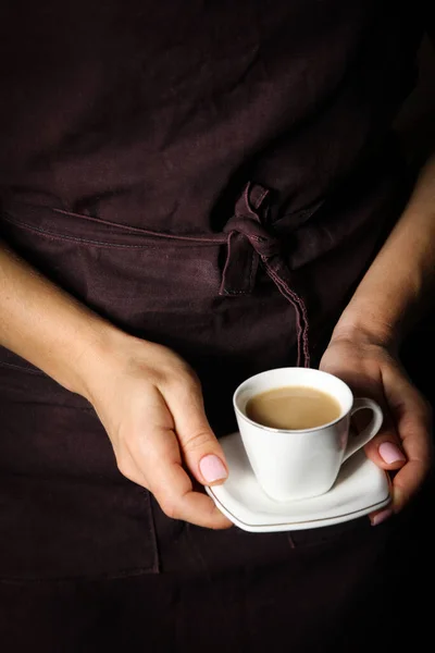 Coffee with cream in a white cup in the hands of a waiter in a black apron. Background image, copy space