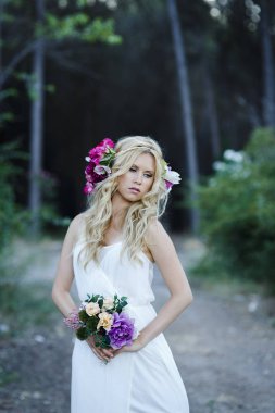 Kırsal kesimde uzun dalgalı saçlı ve mavi gözlü genç sarışın kadınla. Beyaz elbise, çiçek hairband ve çiçek buketi tutan çekici gelin