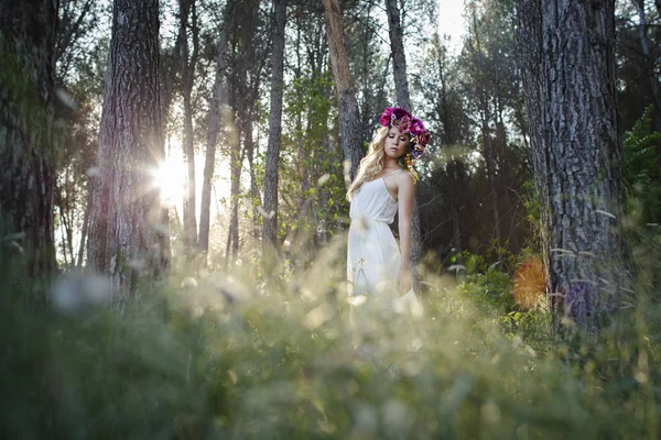 Blond Caucasian woman with wavy hair, posing in romantic style in the forest. Young beautiful bride wearing elegant white dress, flowers and backlight