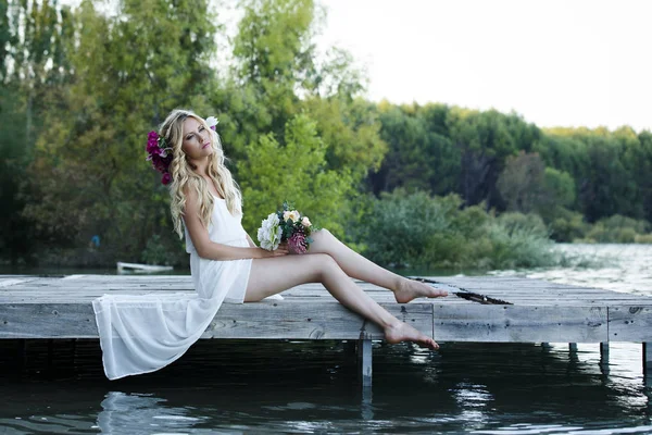 長いウェーブのかかった髪の花の花束を持って魅力的な金髪の女性 湖の横にある桟橋に座って花のヘアバンドを身に着けているロマンチックな花嫁 — ストック写真