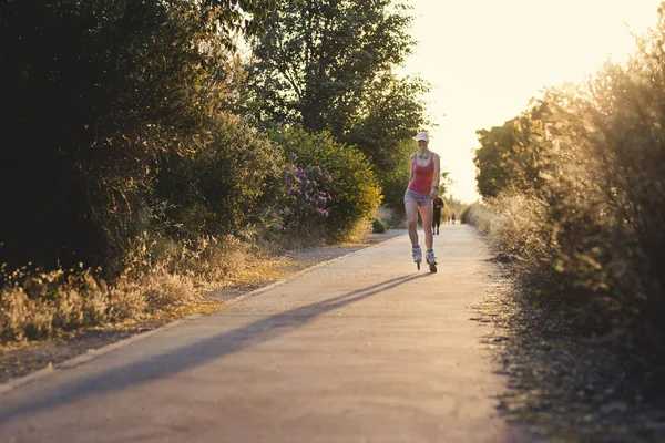 Gün batımında paten kayarken eğlenen genç bir kadın. Soluk pembe şapkalı, kulaklıklı ve spor elbiseli çekici bir kadın egzersiz yapıyor..