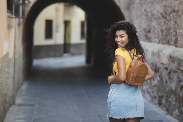 Young mixed woman with curly hair having fun in the urban surroundings. African girl wearing casual clothes and cute backpack smiling and looking back while walking on the street.