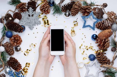 Erkek Noel koniler, kar taneleri ve konfeti ile beyaz arka plan üzerinde akıllı cep telefonu tutan eller. Düz yatıyordu kompozisyon üstten görünüm.