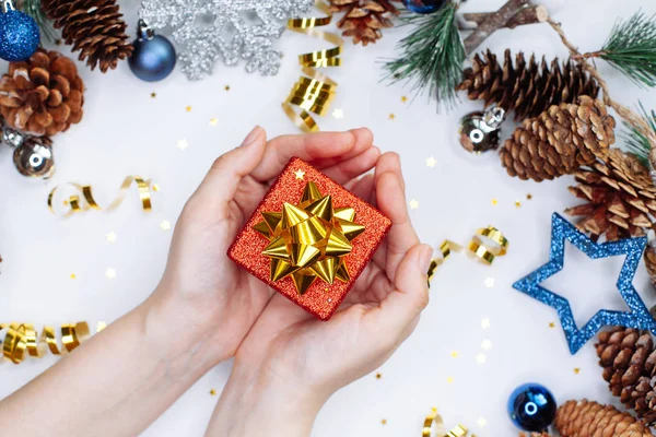 Woman holding a Christmas gift in hands on the background of  fir branches with cones and christmas balls in silver and blue colours