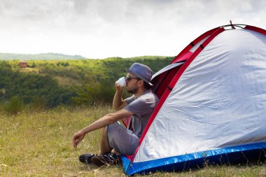 Genç bir turist bir sakal ve kap ile oturan ve onun vaktinizi kamp çadırı içme. Yalnız kamp, açık havada etkinlik doğa yaşam tarzı consept.