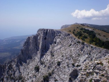Taşlarla kaplı bir manzara. Uçurumun tepesinde parlak yeşil çimenler ve koyu yeşil ağaçlar yetişiyor. Uzakta, dağlar ve vadi sisin içinde görülebilir..