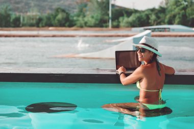 Bikinili genç ince kadın freelancer, hasır şapka ve güneş gözlüğü inşaat dizüstü dizüstü bilgisayar ile yüzme havuzunda oturuyor.