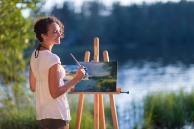 Genç ve güzel bir kadın sanatçı açık havada açık havada bir göl resmi çiziyor.