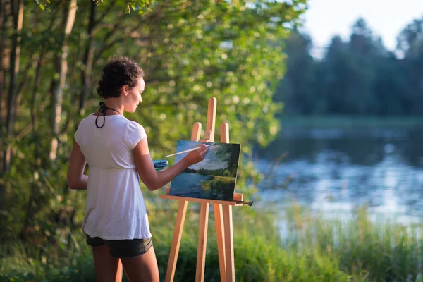 Young pretty woman artist draws paints a picture of lake on open plain air outdoors — Stock Photo, Image