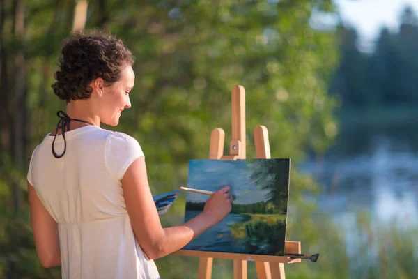 Young pretty woman artist draws paints a picture of lake on open plain air outdoors — Stock Photo, Image