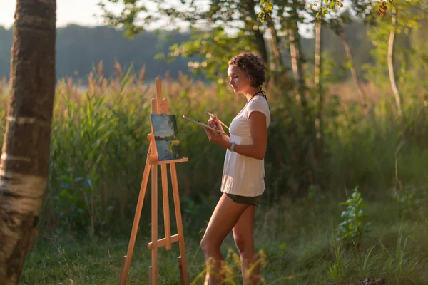 Young pretty woman artist draws paints a picture of lake on open plain air outdoors — Stock Photo, Image