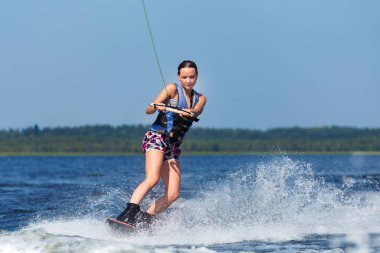 Young pretty slim brunette woman riding wakeboard on wave of motorboat in summer lake clipart
