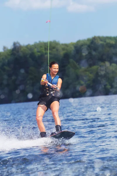 Young pretty slim brunette woman riding wakeboard on wave of motorboat in summer lake — Stock Photo, Image