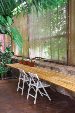 Beyaz sandalyeli ahşap masa ve geniş pencereli saksı bitkileri, bahçenin arka planında yemyeşil, huzurlu bir atmosfer. İş yeri, iş yeri, sera içi ofis, turuncu.