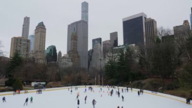İnsanlar, gri ve bulutlu bir günde arkadaşlarıyla Central Park 'taki buz pateni pistini paylaşarak ve arka planda metropolün devasa gökdelenlerini paylaşarak buz patenini seviyorlar.