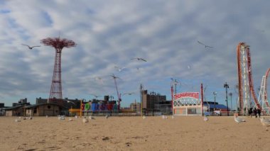 Martılar Coney Adası 'nda Coney Adası' nın arka planında Coney Adası eğlence parkı ve eğlence parkı olan açık gökyüzünde hareket eden beyaz bulutların altında uçuyor ve yürüyorlar.