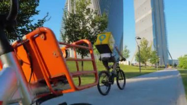 Milan, Haziran 2020 'de bir bisikletli kurye parkın ortasındaki bir caddede hızla geçiyor. Arka planda modern gökdelenler var. Önde park edilmiş bir bisiklet var. Kamera hareketini yukarı eğ