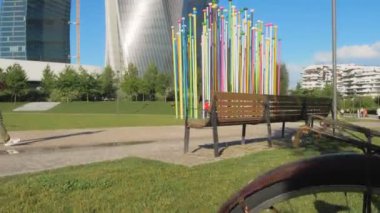 Milan, Haziran 2020, park halindeki bir bisikletten oyuncak bebek kamera hareketi. Adam şehir parkındaki bir sokakta cep telefonuyla konuşuyor. Arka planda modern gökdelenler ve renkli bir sanat kurulumu var.