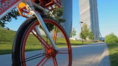 Milan, Haziran 2020 - Milano 'da bir şehir parkında park etmiş bir bisikletle başlayan dikey kamera hareketi çimenleri geçen bir yol ve güneşli bir günde arka planda modern gökdelenler