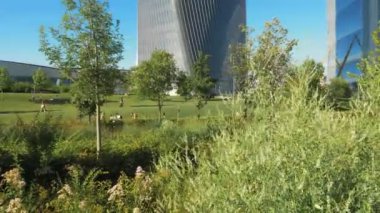 İnsanlar Milano 'daki bir şehir parkında güneşli bir günde yürüyüp boş zamanlarının tadını çıkarıyorlar. Ön planda bir çalı ve arka planda modern gökdelenler. kamera sağa kaydır