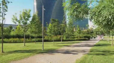 Önünde kumlu bir patika olan şehir parkı ve arka planında gökdelenler. Uzakta güneşli günün tadını çıkaran bir aile. Kamera bir ağacın arkasına geçene kadar sağa kayar.