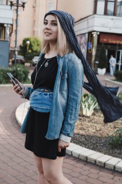 Elinde telefon ve kulaklık ile sokakta duran mavi kemer denim çanta ile genç şık kadın. Gülümseyerek ve müzik dinleme