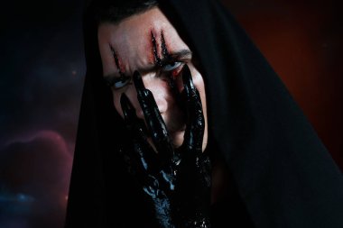 An evil demon hides his face behind his black hand and looking to the camera. Halloween makeup concept clipart