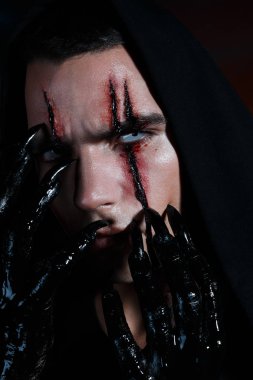 black magician scratches his face with black claws leaving bloody marks on it. Halloween horror concept clipart