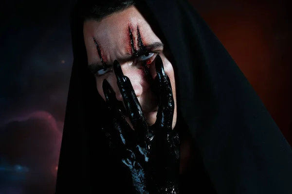 An evil demon hides his face behind his black hand and looking to the camera. Halloween makeup concept