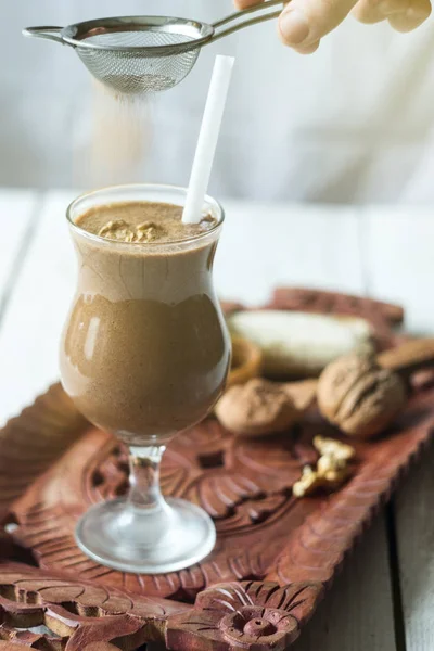 Homemade banana wallnut brown smoothie in a glass on a wooden tray. Hand sprinkles with cinnamon over it. Delicious breakfast