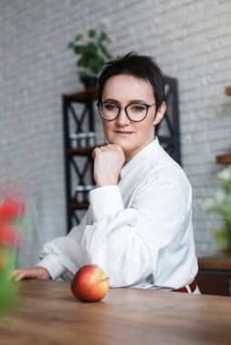 Bardaklı esmer kız mutfakta elmayla oturuyor.