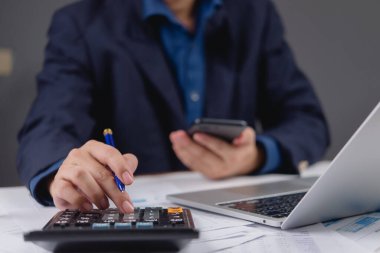 İş adamı, hesap makinesi ve cep telefonu kullanarak finansal verileri hesaplıyor. Belgeler, dizüstü bilgisayarlar ve mali planlama raporlarıyla dolu bir ofis masası. Bütçe maliyet analizi.