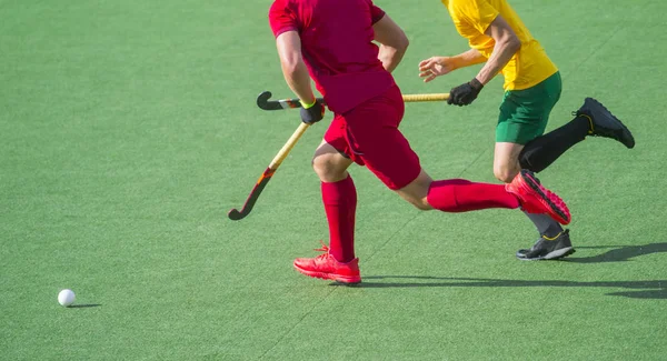 İki çim hokeyi oyuncusu, yapay çimlerde yoğun bir maç sırasında orta sahada top için dövüşüyor.