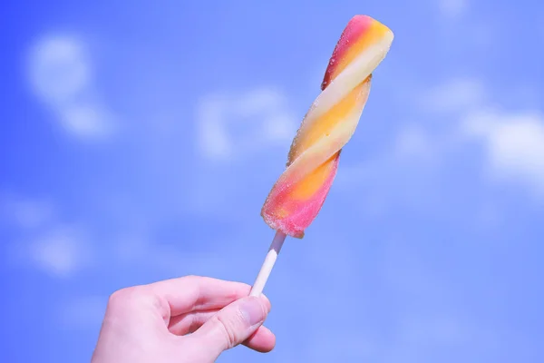 Hand holding orange, yellow and red ice lolly pop with Summer blue and clouds sky background