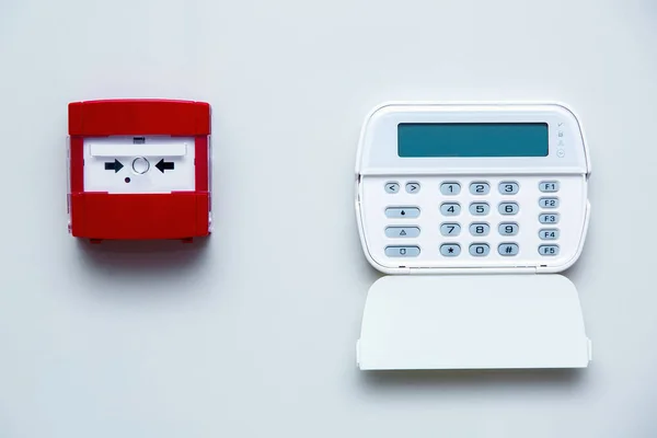 Fire safety button in a red box on a gray wall near a white alarm control panel with security password input buttons, front view on a bright wall indoors.