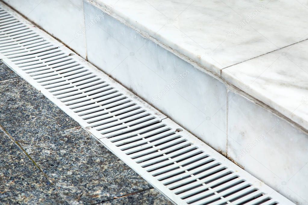 The gutter of the drainage system on the sidewalk, close-up metal grille light color on the background of marble tiles.