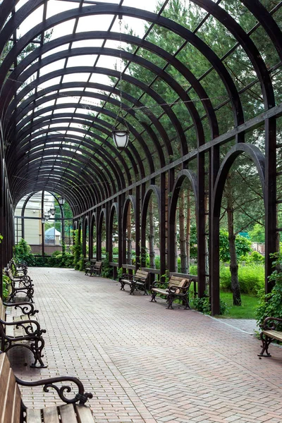 Arched gazebo with wooden arch with retro lanterns and benches with forged legs and wooden seat mats, a place to walk with the sidewalk.