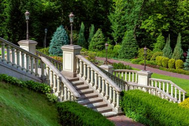 bir taş merdiven korkuluk ve korkuluklar üzerinde kaideleri ile yüklenir demir fener ayağı hedges çalılar ve ağaçlar şeklinde yeşillik ve peyzaj tasarımı ile bir parkta dövme fener ile.
