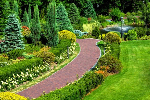 ランドス ケープ デザインと様々 なブッシュ植物や木 庭園での散歩の通路の上から見る庭の赤い舗装通路 — ストック写真